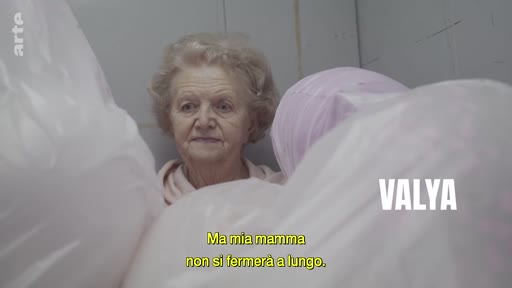 Portrait de Valya, une femme âgée, filmée dans un environnement intérieur avec des sacs en plastique au premier plan. Une incrustation à l'écran indique son prénom et une sous-titre en italien exprime une réflexion sur sa mère.