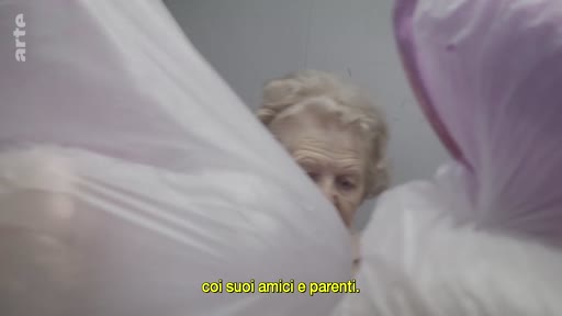 Une femme âgée est filmée en gros plan, partiellement cachée par des sacs en plastique blancs, dans une séquence évoquant ses relations avec ses amis et parents.