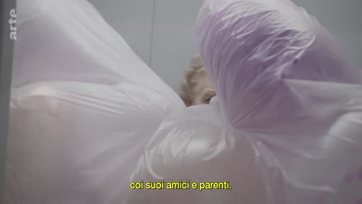 Une personne, dont le visage est partiellement visible, se trouve derrière des sacs en plastique, dans une scène évoquant des relations sociales ou familiales.