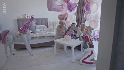 Une famille ukrainienne célèbre un anniversaire dans une chambre décorée de ballons à Kyiv, avec un grand ballon en forme de licorne et un gâteau sur une petite table. Les membres de la famille prennent un selfie ensemble pour immortaliser ce moment festif.