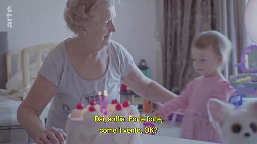 Une grand-mère et sa petite-fille célèbrent un anniversaire à Kyiv, en Ukraine. La grand-mère encourage l'enfant à souffler les bougies sur un gâteau, dans un contexte de vie familiale malgré le conflit en cours.