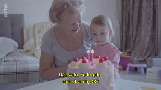 Une grand-mère aide une jeune enfant à souffler les bougies sur un gâteau d'anniversaire lors d'une célébration familiale à Kyiv, en Ukraine. La scène montre un moment intime et chaleureux dans un contexte de vie quotidienne en temps de guerre.