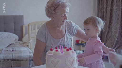 Une femme âgée et une jeune enfant célèbrent un anniversaire devant un gâteau rose décoré de cœurs à Kyiv, en Ukraine. Cette scène illustre la vie quotidienne et les moments familiaux dans un contexte de conflit.