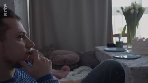 Un jeune homme à l'air préoccupé, la main sur le menton, est assis dans un salon. Cette scène illustre une atmosphère d'inquiétude ou de réflexion profonde dans un contexte domestique.