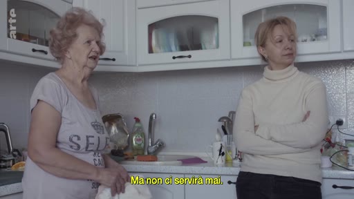Deux femmes discutent dans une cuisine, exprimant leurs inquiétudes face à une menace imminente. L'une d'elles porte un t-shirt avec l'inscription 'SELF', tandis que l'autre est vêtue d'un col roulé beige.