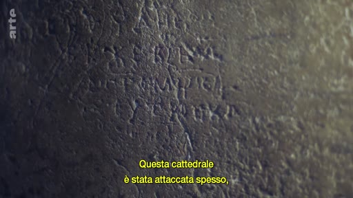 Gros plan sur une inscription ancienne gravée dans la pierre de la cathédrale Sainte-Sophie, témoignant de son histoire mouvementée. Le texte à l'écran souligne que cette cathédrale a été attaquée souvent.