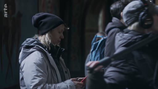 Une équipe de tournage, incluant une femme portant un bonnet noir et une veste grise, filme à l'intérieur de la cathédrale Sainte-Sophie. Le cadre montre les coulisses de la production audiovisuelle sur ce site historique.