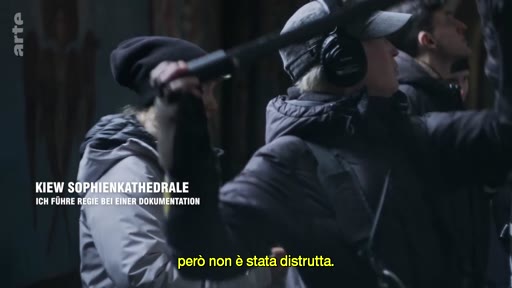Une équipe de tournage, équipée de casques audio et de perches micro, filme à l'intérieur de la cathédrale Sainte-Sophie à Kiev, en Ukraine. Le texte à l'écran indique le lieu et le contexte de la réalisation d'un documentaire.