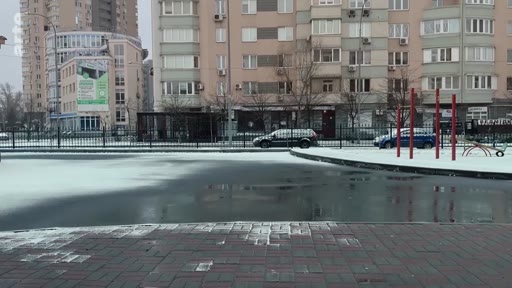 Vue d'ensemble d'une place urbaine enneigée avec une patinoire extérieure, entourée d'immeubles résidentiels en hiver. Le paysage urbain montre des conditions hivernales avec de la neige au sol et une surface glacée.