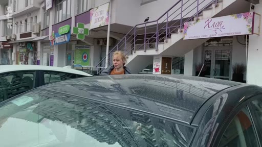 Une femme blonde marche devant des vitrines de magasins avec des enseignes en ukrainien, incluant une pharmacie et une boutique de décoration, dans une rue urbaine en Ukraine.