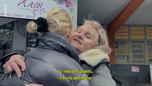 Deux femmes se serrent dans les bras lors d'un moment d'adieu émouvant, avec un sous-titre en italien indiquant 'Ci vediamo presto, tra una settimana'. La scène se déroule devant une vitrine de magasin avec des inscriptions en ukrainien.