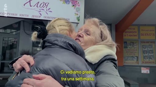 Deux femmes se serrent dans les bras lors d'un moment d'adieu émouvant devant une boutique nommée 'ХЛОЯ' en Ukraine. La scène illustre un départ ou une séparation temporaire, avec une légende en italien indiquant 'Ci vediamo presto, tra una settimana'.