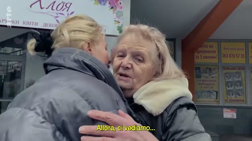 Une femme âgée et une femme plus jeune se font une accolade émouvante dans la rue, marquant un moment de séparation ou de départ. La scène se déroule devant une vitrine de magasin avec des inscriptions en ukrainien.