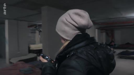 Une personne portant un bonnet et une doudoune noire se trouve dans un abri souterrain aménagé, tenant une télécommande à la main. L'image illustre les conditions de vie dans un refuge, probablement en période de conflit.