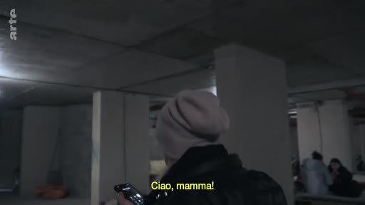 Une personne portant un bonnet communique par téléphone dans un abri souterrain sombre, probablement en Ukraine, alors qu'elle dit 'Ciao, mamma!' en sous-titre.