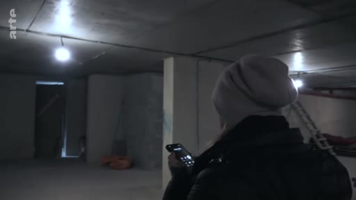 Une personne portant un bonnet et une veste noire consulte son téléphone portable à l'intérieur d'un abri souterrain sombre, illustrant les conditions de vie dans les refuges.