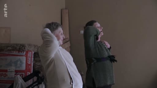 Deux femmes âgées, vêtues de vêtements décontractés, regardent attentivement vers l'extérieur ou vers un point focal hors champ dans une pièce intérieure sombre, illustrant une scène de réflexion sur le conflit.