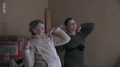 Deux femmes âgées réalisent des mouvements de gymnastique ou d'étirement dans un intérieur simple, illustrant le quotidien ou la résilience de civils dans un contexte de conflit.