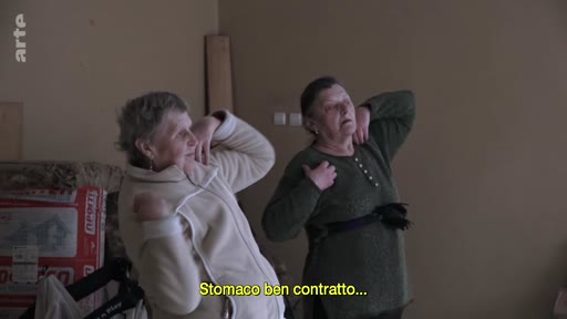 Deux femmes âgées pratiquent des exercices de gymnastique ou de rééducation dans un intérieur, avec une légende en italien à l'écran mentionnant le maintien du ventre contracté.