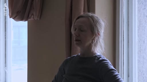 Portrait d'une femme blonde, vue de profil, se tenant debout devant une fenêtre dans une pièce à la lumière tamisée. Cette image illustre une séquence de réflexion personnelle ou de témoignage dans le cadre d'un reportage.