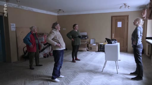 Quatre personnes, dont trois femmes âgées et une instructrice, effectuent des exercices physiques dans une salle dépouillée, probablement dans le cadre d'un programme de soutien ou de rééducation en Ukraine.