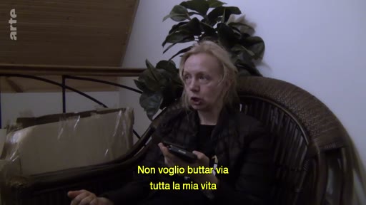 Une femme blonde s'exprime face caméra lors d'une interview, avec des sous-titres en italien affichés à l'écran. Elle discute de son parcours de vie, probablement dans le cadre d'un témoignage sur l'exil ou la condition de réfugié.