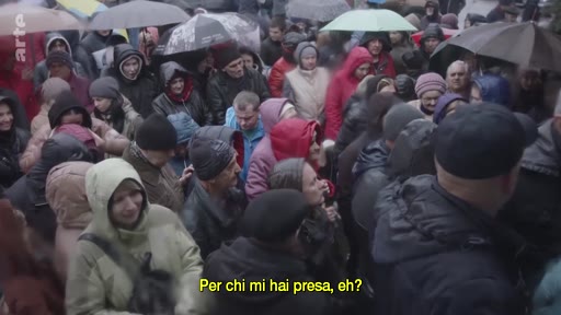Une foule dense de personnes, dont beaucoup portent des parapluies, se rassemble dans un contexte de crise migratoire. Une femme est interviewée au milieu de la foule, avec une légende en italien affichée à l'écran.