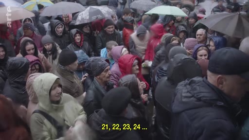 Une foule dense de personnes, dont beaucoup portent des capuches et utilisent des parapluies, se rassemble dans un espace extérieur par temps pluvieux. L'image illustre une situation de crise humanitaire ou de déplacement de population.