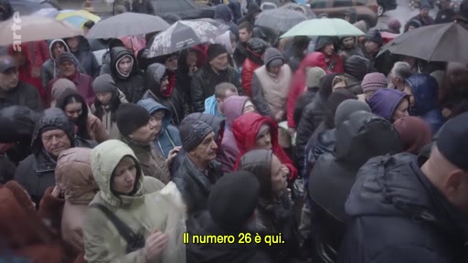 Une foule dense de personnes, principalement des femmes et des personnes âgées, attend sous la pluie, probablement dans le cadre d'une procédure d'enregistrement ou d'aide humanitaire. Le sous-titre indique 'Il numero 26 è qui', suggérant une organisation par numéros pour la gestion des réfugiés.