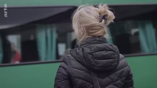 Une femme blonde vue de dos, portant une doudoune noire, se tient devant un bus de couleur verte lors d'un reportage sur les réfugiés.