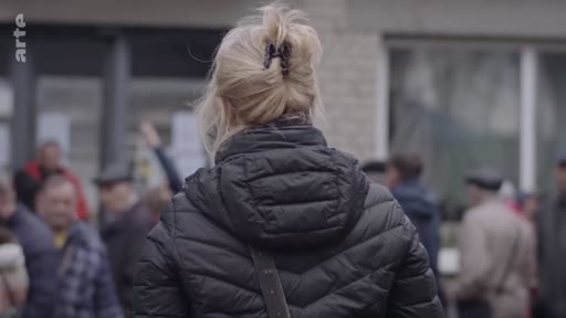 Une femme blonde vue de dos, portant une doudoune noire, se trouve au milieu d'un groupe de personnes lors d'un rassemblement en extérieur.