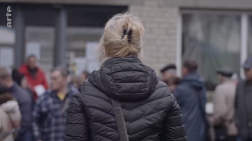 Une femme blonde vue de dos, portant une doudoune noire, se tient au milieu d'une foule de personnes dans un espace extérieur. La scène évoque une situation de rassemblement ou d'attente, potentiellement liée à une crise migratoire ou humanitaire.