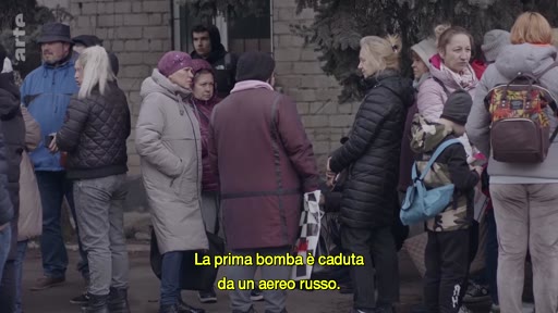 Un groupe de civils, incluant des femmes et des enfants, se rassemble à l'extérieur dans un contexte de conflit. Le sous-titre italien indique que la première bombe a été larguée par un avion russe, soulignant la situation de crise humanitaire et de déplacement de population.