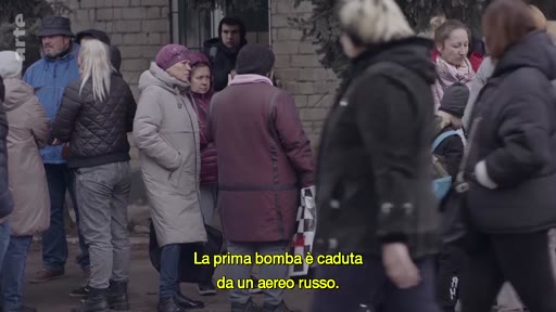 Un groupe de civils, incluant des femmes et des hommes, se tient dans une rue, illustrant la situation des personnes déplacées en Ukraine. Une incrustation de texte en italien indique que la première bombe a été larguée par un avion russe.