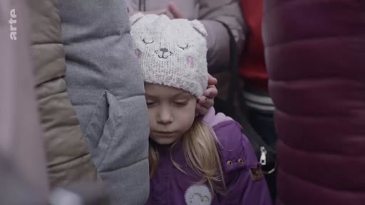 Une jeune enfant blonde portant un bonnet à oreilles en tricot semble épuisée alors qu'elle est entourée par une foule de personnes, illustrant la détresse des civils en situation de déplacement ou de crise humanitaire.