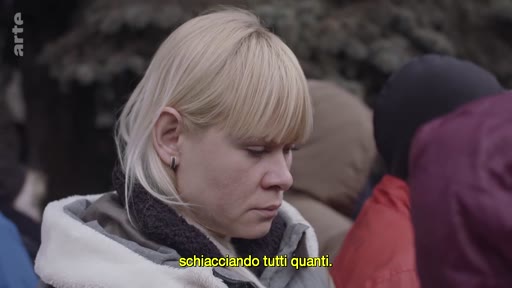 Une femme blonde au regard baissé, visiblement pensive ou triste, se trouve au milieu d'une foule lors d'un événement en extérieur. La scène, sous-titrée en italien, évoque un contexte de déplacement ou de crise humanitaire.
