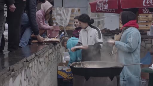 Des bénévoles, dont une femme en blouse bleue, préparent un repas chaud dans une grande marmite en extérieur pour des personnes déplacées. La scène montre une distribution alimentaire organisée dans un contexte de crise humanitaire.