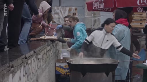 Des bénévoles préparent et distribuent de la nourriture dans une grande marmite pour des personnes déplacées, dans un contexte d'aide humanitaire. La scène montre une organisation de secours sur le terrain avec des bénévoles portant des gants et des vêtements de protection.