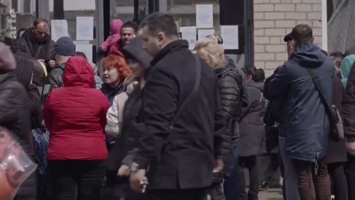 Des civils font la queue devant un bâtiment administratif ou un centre d'aide à Marioupol, en Ukraine, dans un contexte de crise humanitaire liée au conflit. La scène montre des personnes attendant patiemment devant des affiches placardées sur une porte vitrée.