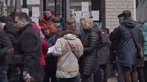 Des civils font la queue devant un bâtiment à Marioupol, en Ukraine, pour recevoir de l'aide humanitaire. Une personne porte une veste avec l'inscription 'DRC' (Danish Refugee Council), témoignant de la présence d'organisations internationales dans la ville dévastée.