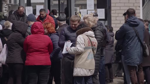 Des civils ukrainiens font la queue devant un bâtiment à Marioupol pour obtenir de l'aide ou des documents, dans le contexte de la guerre en Ukraine. Une personne porte un gilet avec l'inscription 'DRC', suggérant une aide humanitaire sur place.