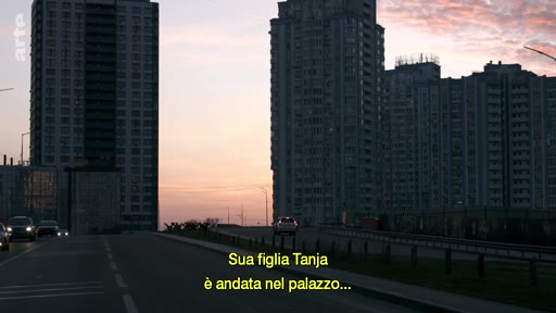 Vue extérieure d'une route urbaine entourée de grands immeubles résidentiels au coucher du soleil, accompagnée d'un sous-titre en italien mentionnant la fille Tanja.