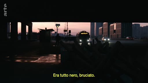 Vue urbaine en contre-plongée montrant un bus et des voitures circulant sur une route au crépuscule, illustrant un contexte de voyage ou de déplacement dans un environnement urbain. L'image est accompagnée d'un sous-titre en italien évoquant un paysage sombre et brûlé.