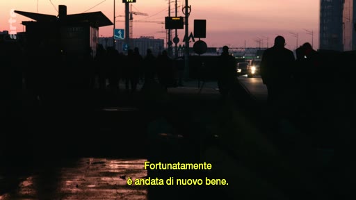 Des silhouettes de personnes marchent dans une rue urbaine au crépuscule, illustrant le thème des parcours de réfugiés et de la survie. Le sous-titre indique 'Fortunatamente è andata di nuovo bene'.