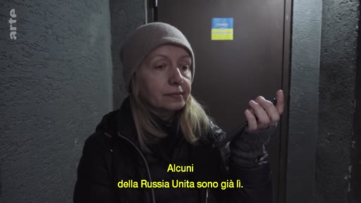 Une femme portant un bonnet s'exprime lors d'une interview dans un espace clos, possiblement un abri, avec un drapeau ukrainien visible en arrière-plan. Le sous-titre mentionne le parti Russie Unie dans le contexte de la guerre en Ukraine.