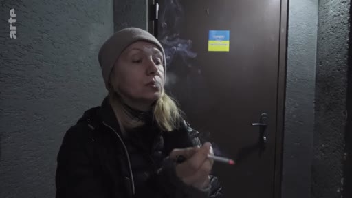 Une femme portant un bonnet fume une cigarette dans un espace sombre, devant une porte sur laquelle est apposé un petit drapeau de l'Ukraine. Cette image illustre les conditions de vie précaires et le quotidien des civils dans le contexte de la guerre en Ukraine.