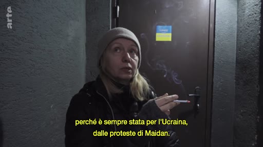 Une femme portant un bonnet s'exprime lors d'une interview, une cigarette à la main, devant une porte sur laquelle est apposé un petit drapeau de l'Ukraine. Le sous-titre évoque son engagement pour l'Ukraine depuis les manifestations de Maïdan.