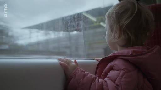 Un jeune enfant, vu de profil, regarde par la fenêtre d'un train en mouvement, observant le paysage défiler. L'ambiance est calme et contemplative lors d'un voyage en train.