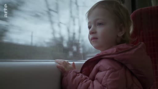 Une jeune enfant blonde, vêtue d'une doudoune rose, observe le paysage défilant par la fenêtre d'un train. Cette image illustre un voyage en train, le transport ferroviaire ou le quotidien des familles en déplacement.