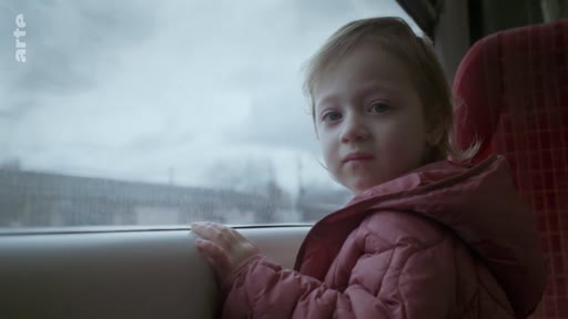 Une jeune enfant, vêtue d'une doudoune rose, observe le paysage défilant par la fenêtre d'un train en mouvement. Cette scène illustre le voyage et l'enfance dans un contexte de transport ferroviaire.